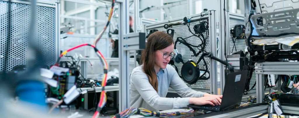 A female engineer working on a laptop in an industrial setting, surrounded by electronic equipment, is focused on her task. Based on the provided description, it's not possible to determine specific SEO keywords due to